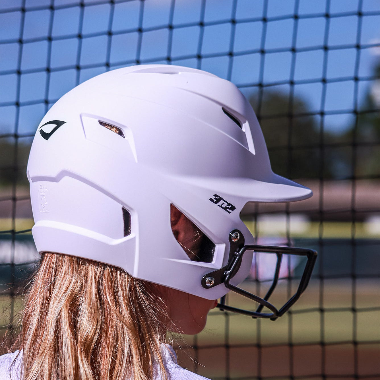 Person wearing a white softball helmet with a black face guard against a netted background